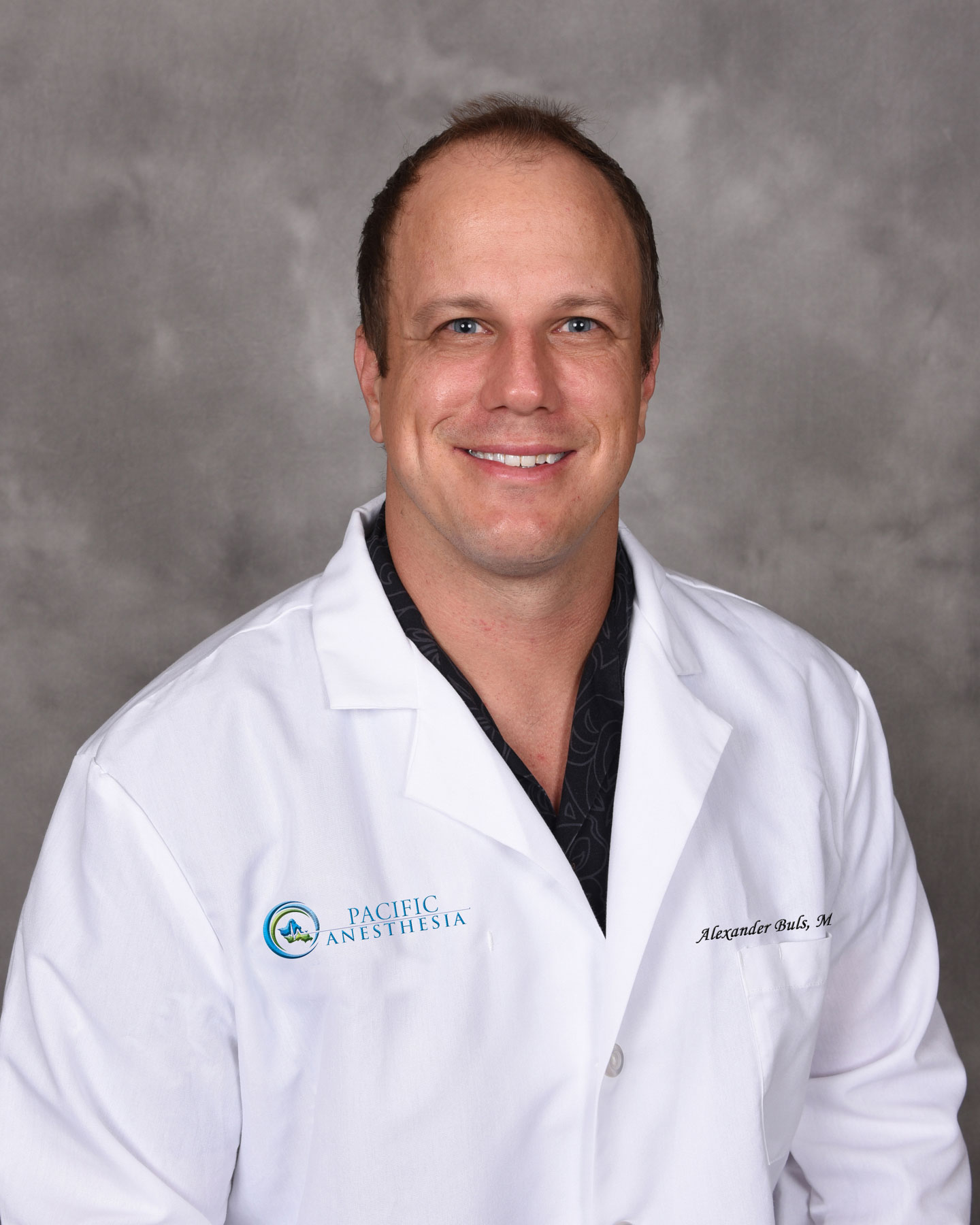 Smiling man wearing a white Pacific Anesthesia coat with black shirt underneath, standing against a gray backdrop.