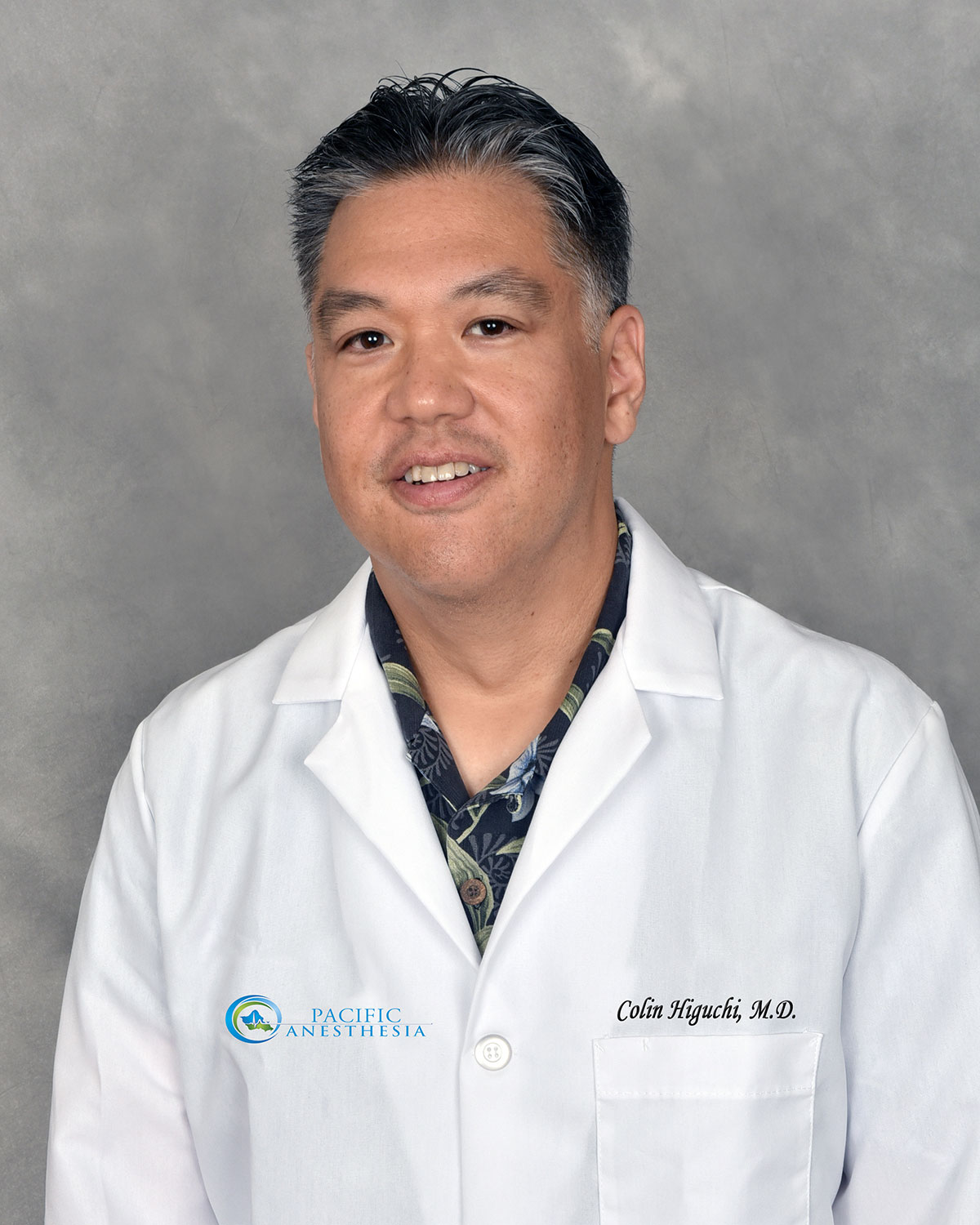 Portrait of a man wearing a white lab coat with 'Pacific Anesthesia' logo and 'Colin Higuchi, M.D.' embroidered on the chest, against a grey background.
