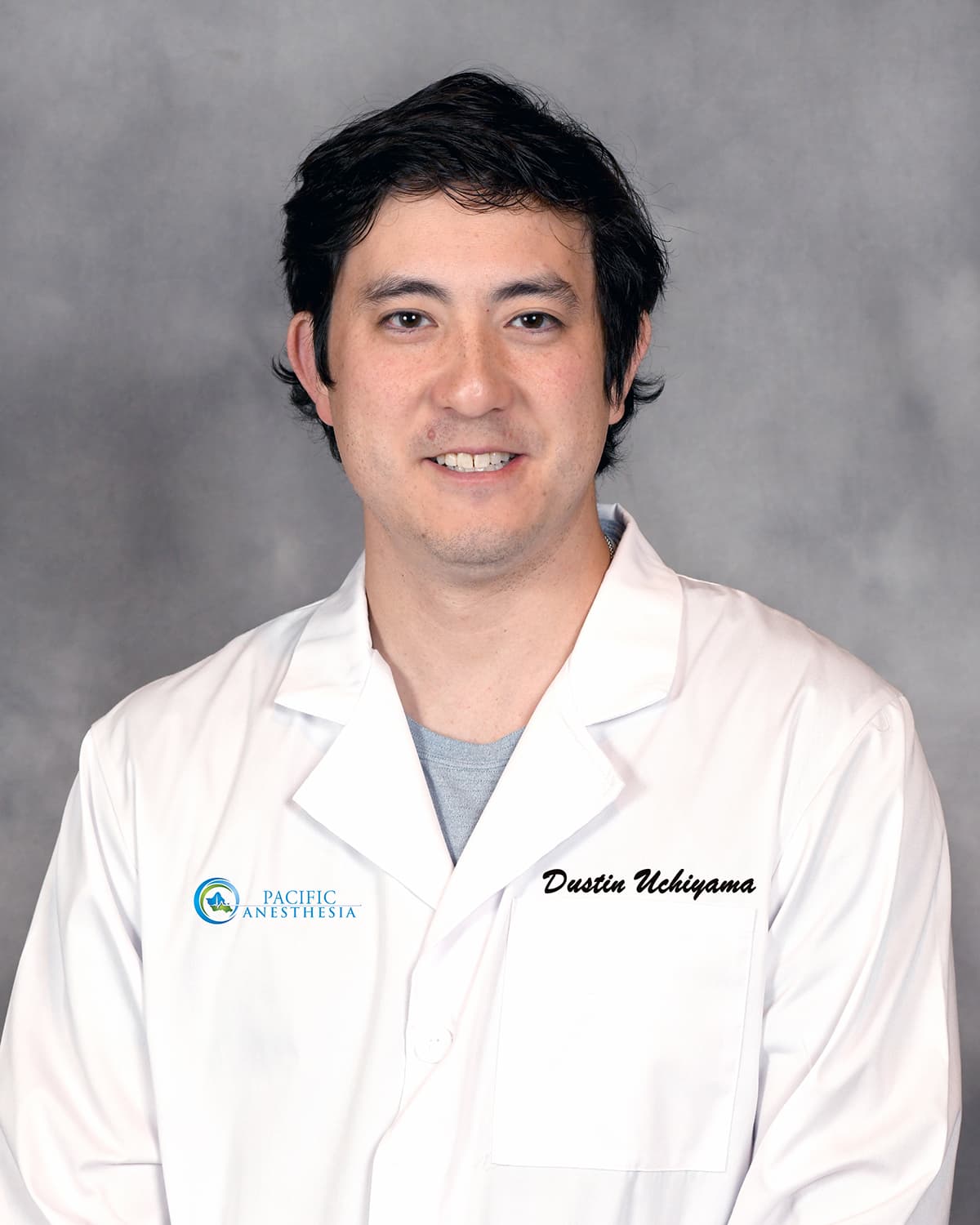 Portrait of a man with dark hair wearing a white lab coat over a gray shirt against a gray background.