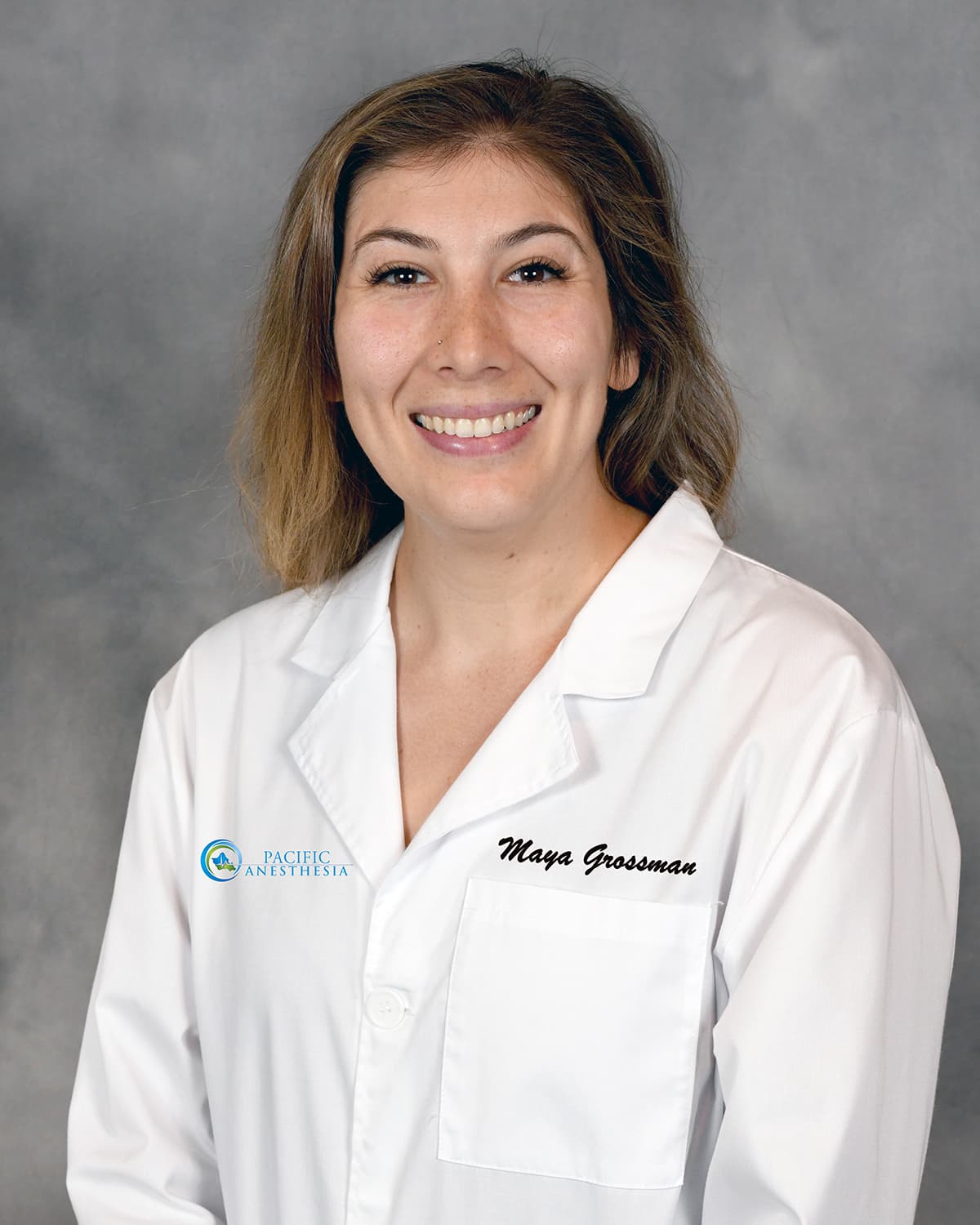 Smiling woman with shoulder-length hair wearing a white lab coat with 'Maya Grossman' and 'Pacific Anesthesia' logos.