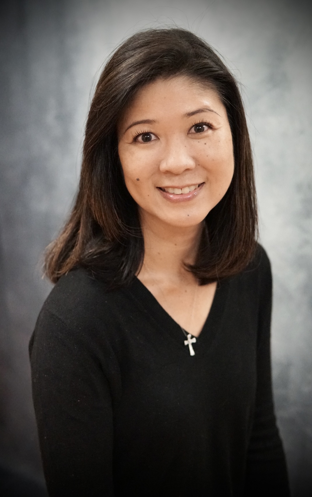Smiling woman with shoulder-length dark hair wearing a black top and a cross necklace against a neutral gray background.