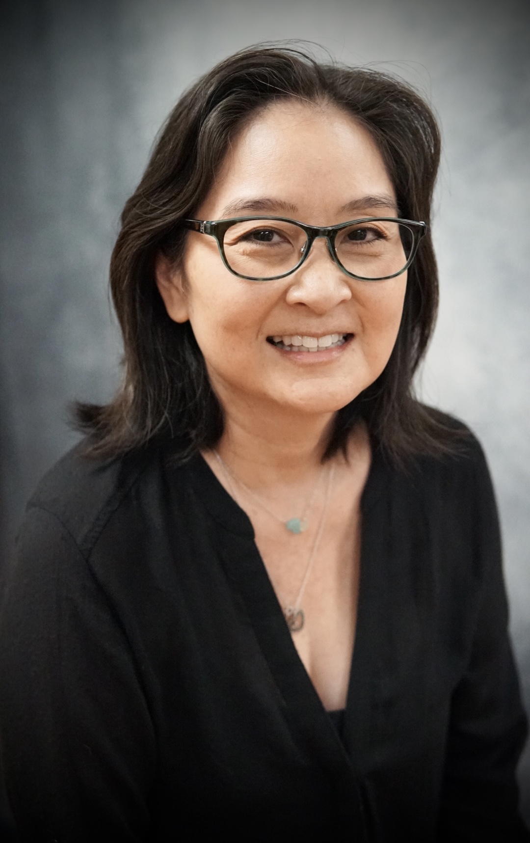 Smiling woman with shoulder-length dark hair wearing glasses and a black top against a gray background.