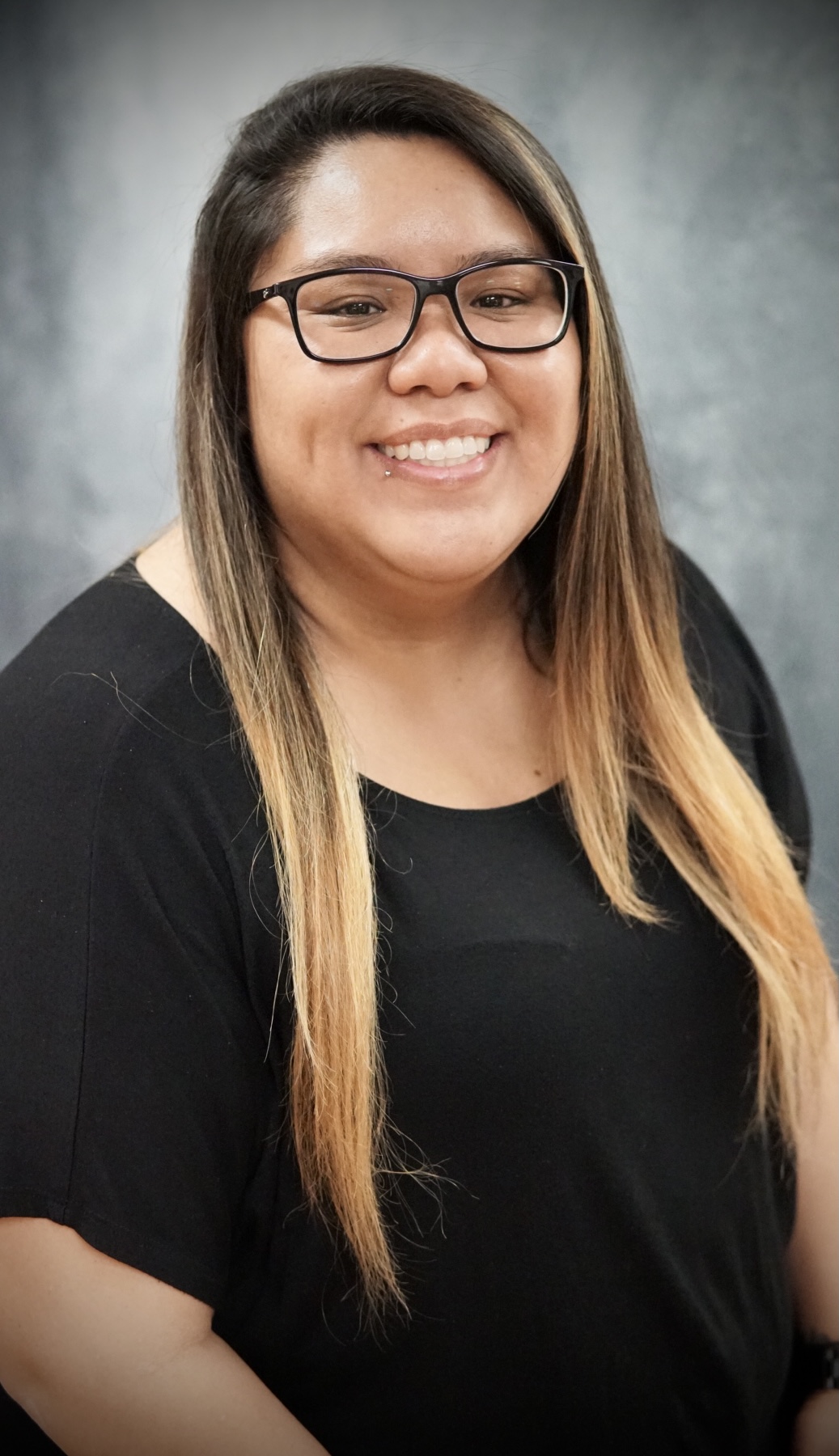 Smiling woman with long ombre hair wearing black glasses and a black top against a gray backdrop.
