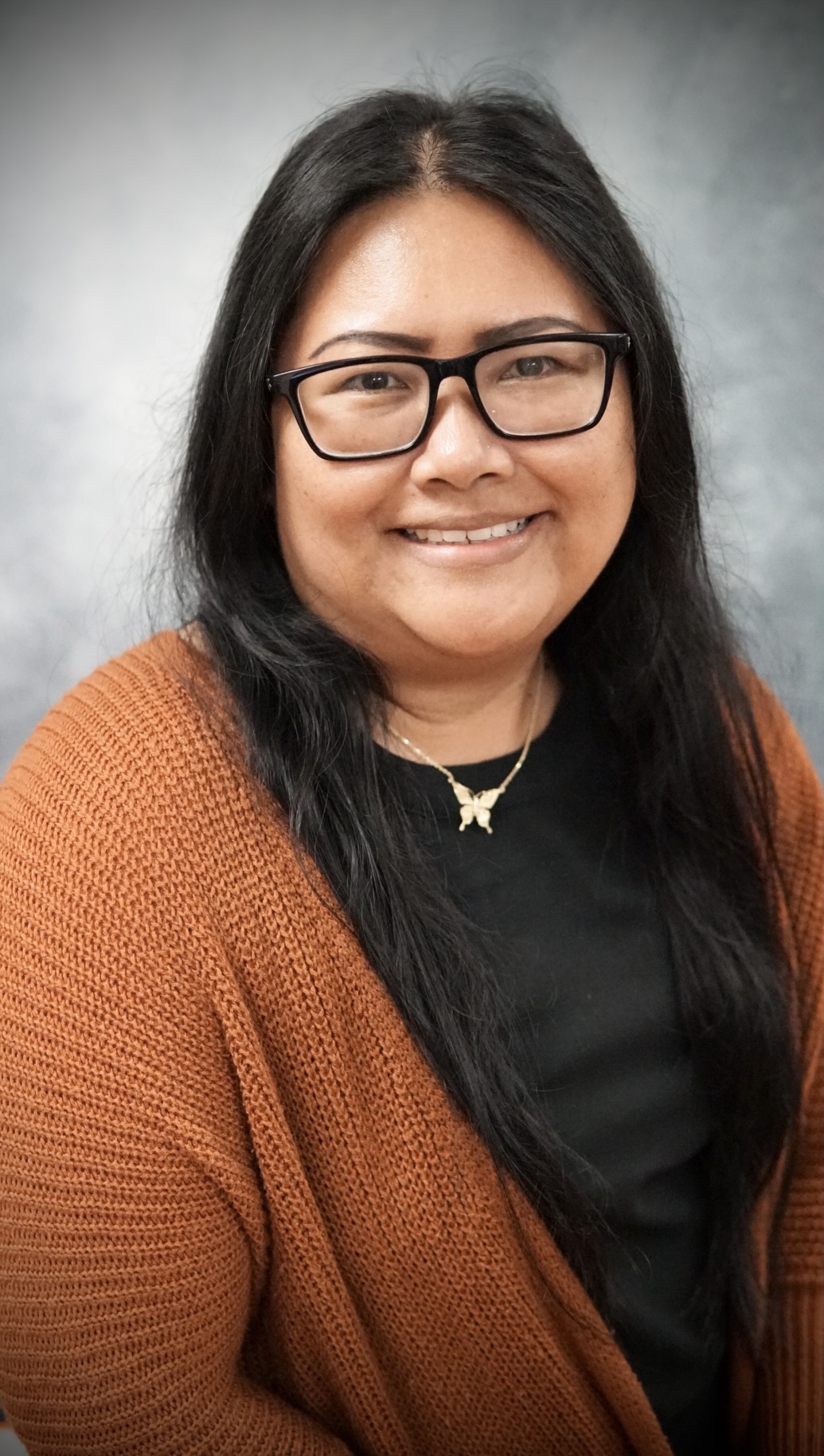 Smiling woman with long black hair, glasses, a butterfly necklace, black top, and rust-colored knit cardigan.