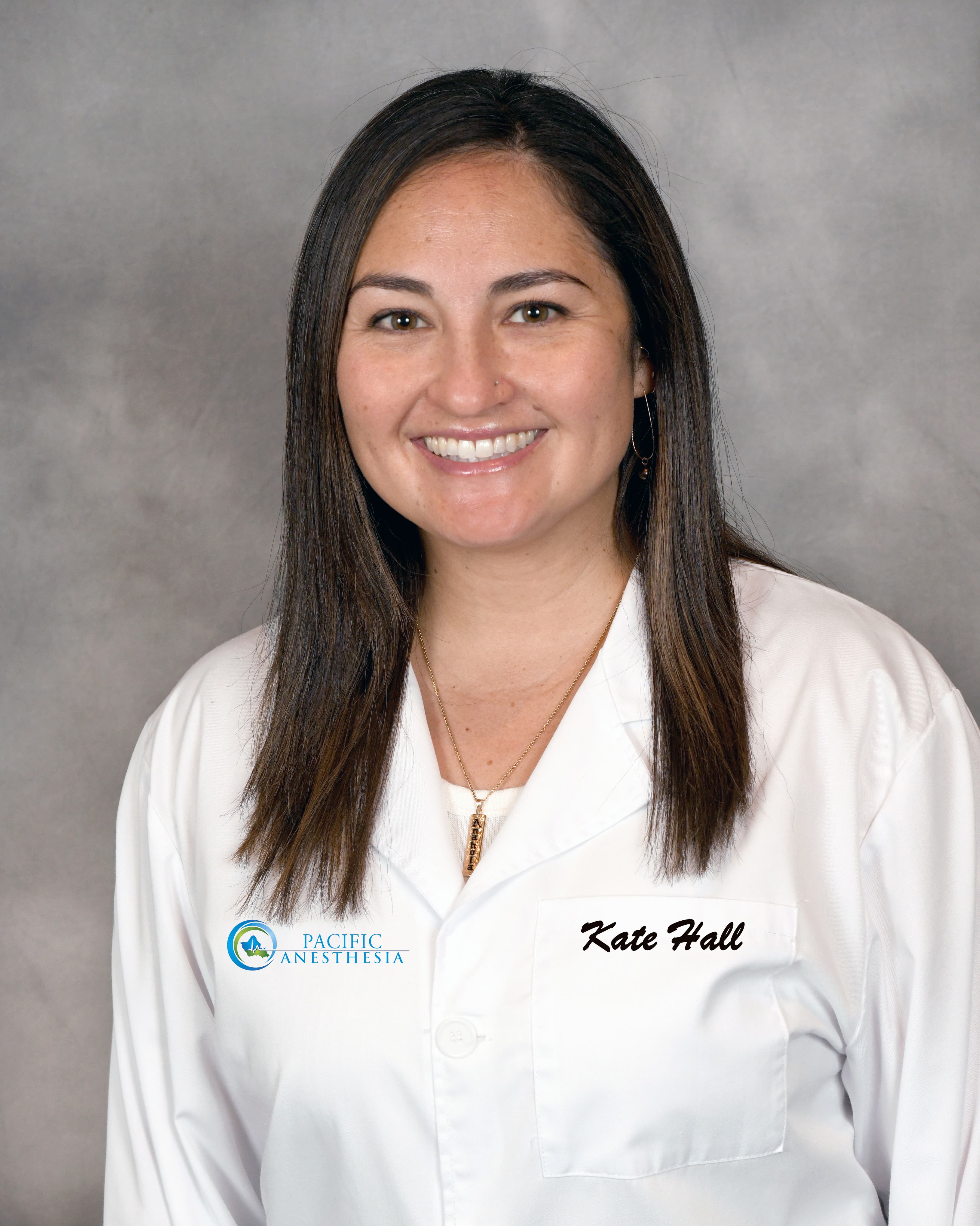 Smiling woman with long dark hair wearing a white medical coat with 'Pacific Anesthesia' logo and name tag.