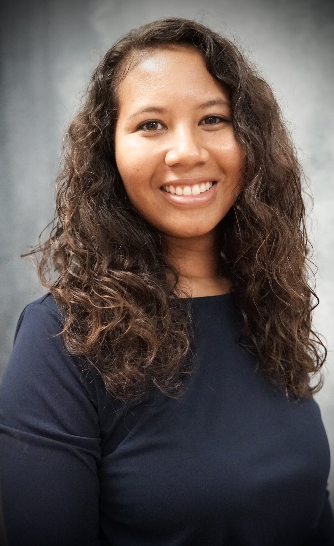 Smiling woman with long curly hair wearing a navy blue top against a gray blurred background.