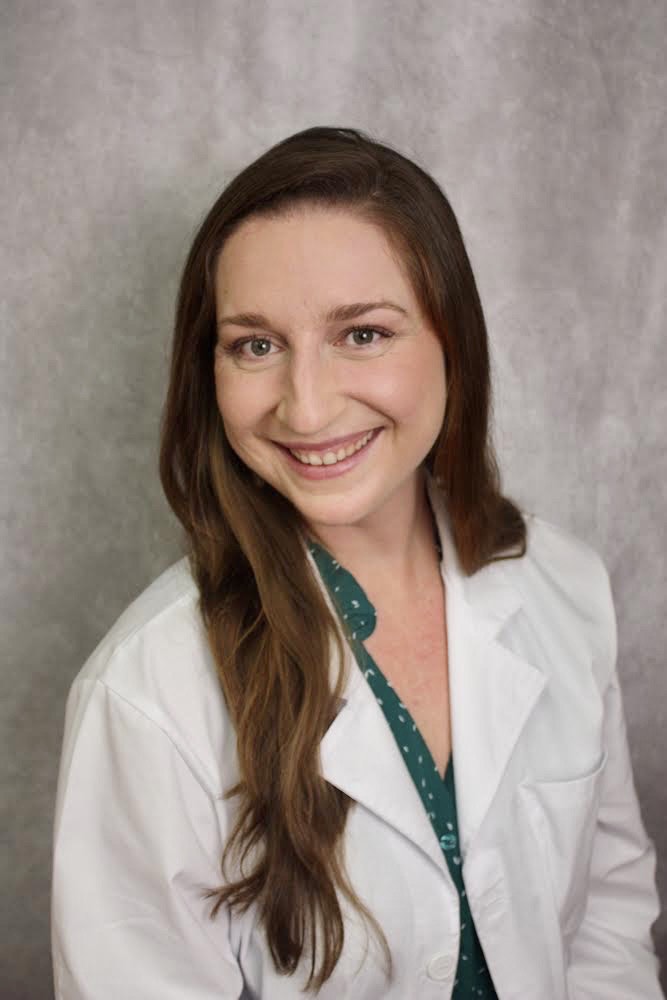 Smiling woman with long brown hair wearing a white lab coat over a green blouse, posed against a gray background.