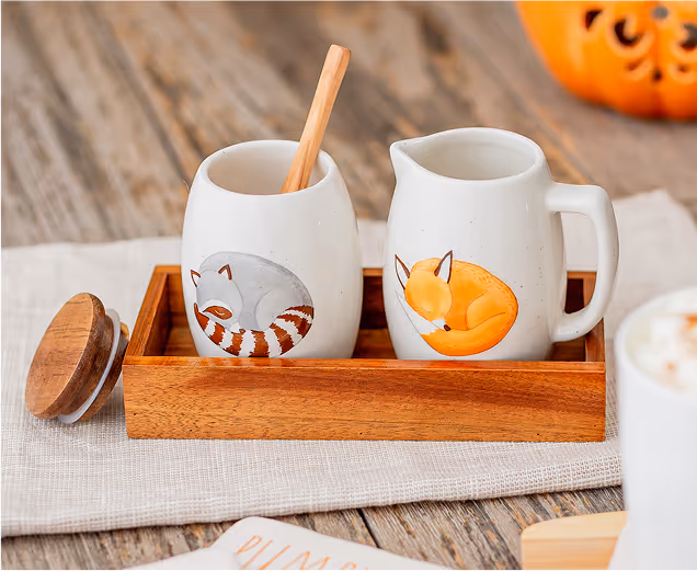 Two ceramic creamers with fox and raccoon illustrations placed on a wooden tray on a rustic table.