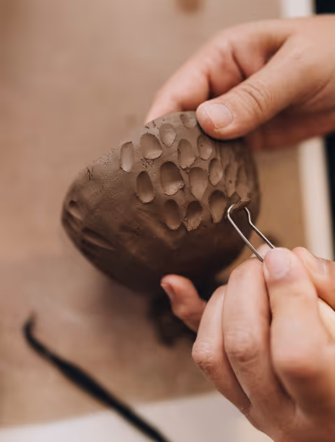 Hands sculpting a small brown clay pot with a binder clip to create patterns.