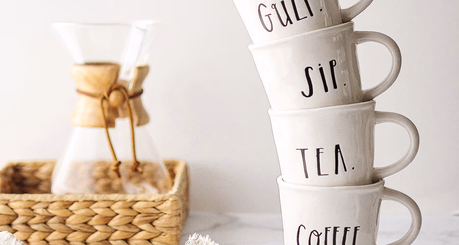 Stack of four white ceramic mugs with words 'GULP.', 'SIP.', 'TEA.', and 'COFFEE.' printed in black, next to a glass coffee brewer in a woven basket.