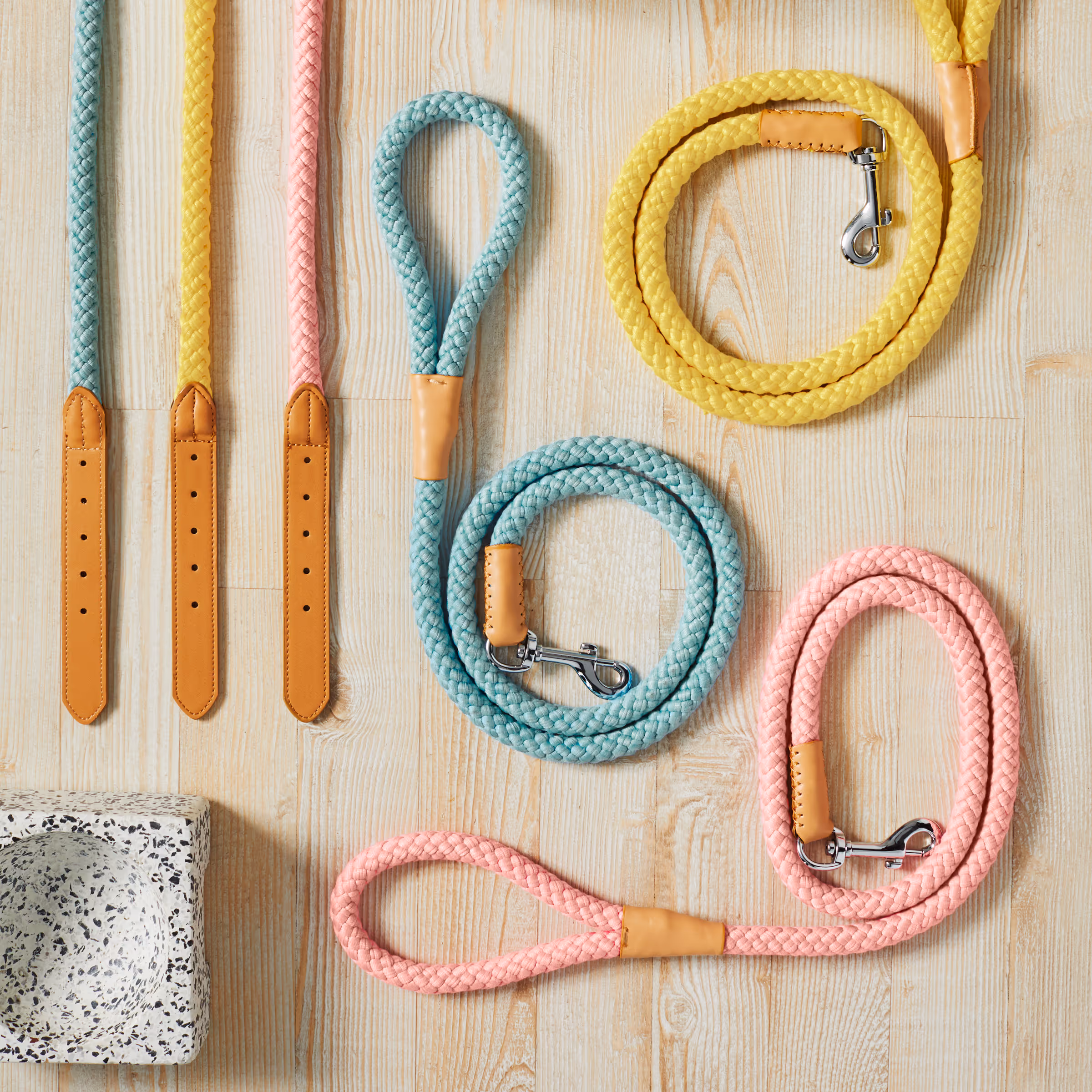 Coiled pastel-colored braided dog leashes with leather handles and metal clasps on a light wooden surface next to a terrazzo dog bowl.