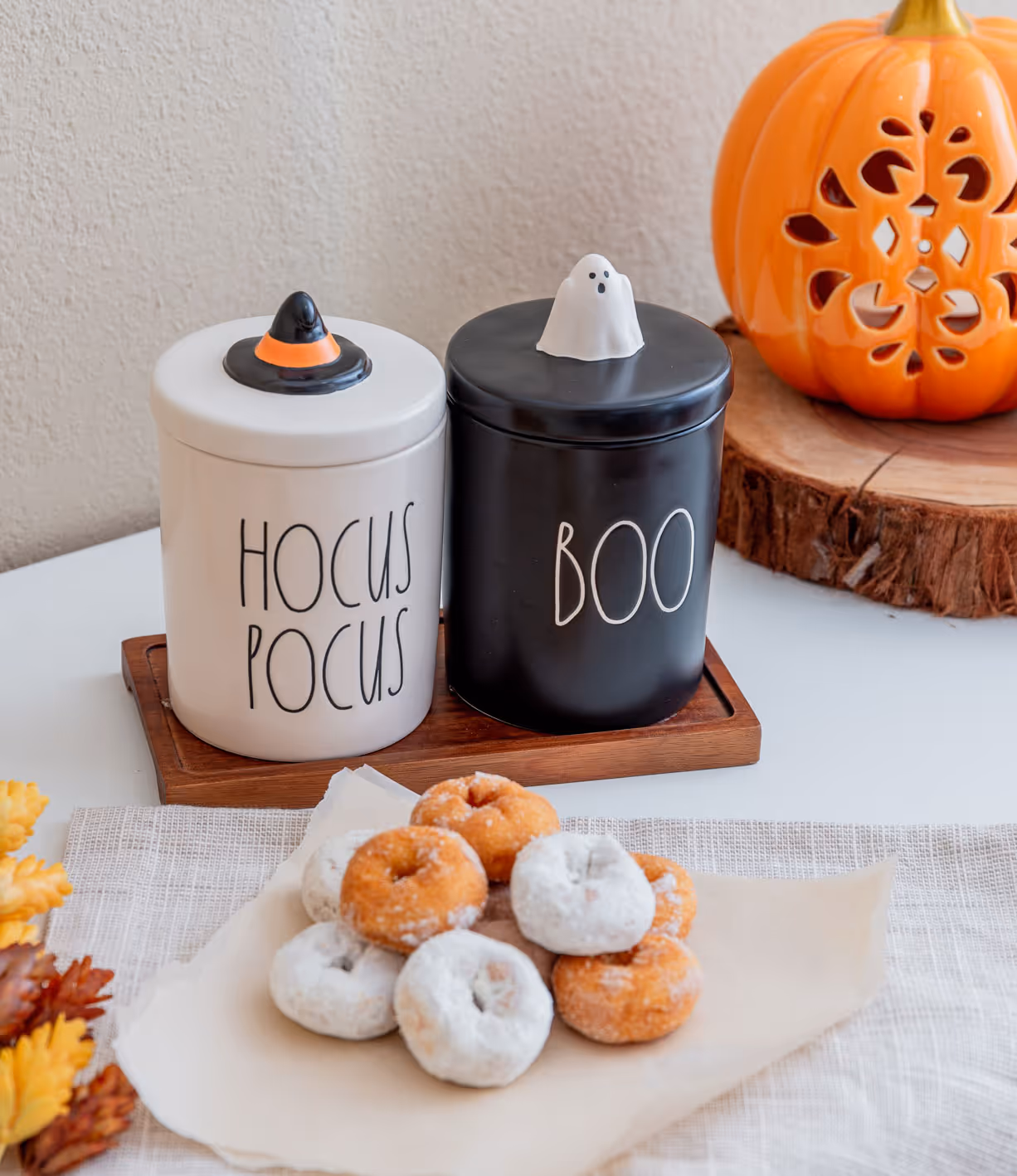 White container labeled 'Hocus Pocus' with a witch hat lid and black container labeled 'Boo' with a ghost lid, next to powdered and plain mini donuts on parchment paper, and a decorative carved pumpkin.