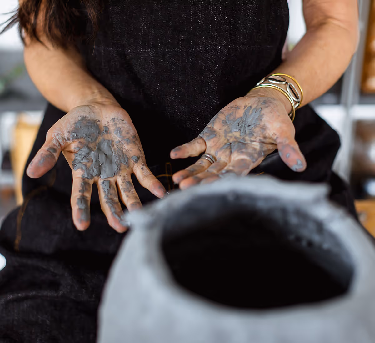 Person showing hands covered in wet clay with a pottery wheel in the foreground.