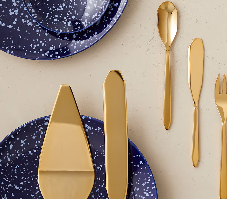 Close-up of gold-colored cutlery set including a spoon, knife, fork, and two serving utensils next to speckled blue plates on a beige surface.
