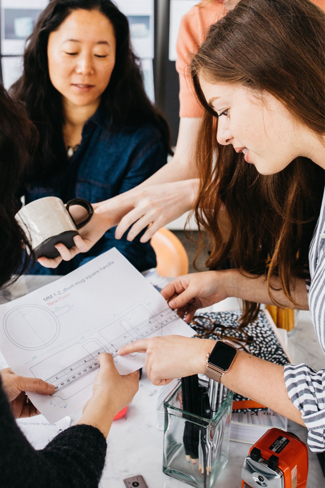 Three women collaborating over a paper with a ruler, one holding a ceramic mug, at a desk with pencils and a sharpener.