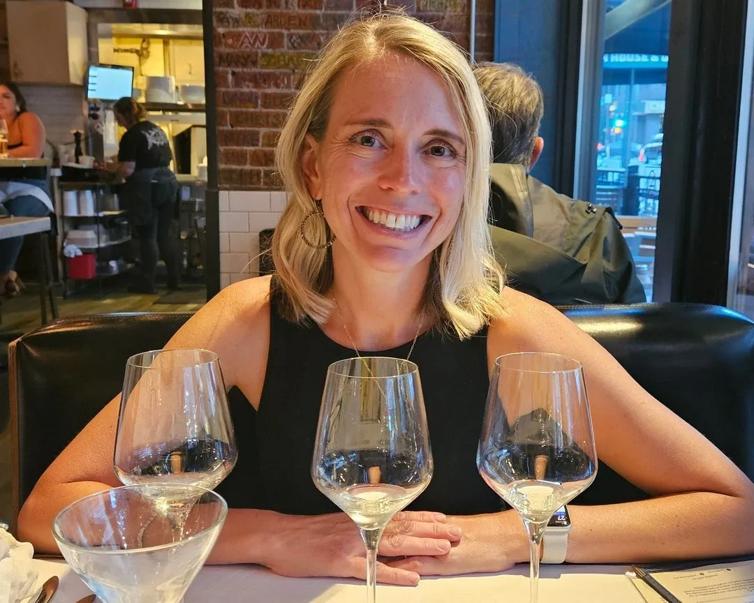 Diana G., from Denver, Colorado, seated at a restaurant with wine glasses in front of her, looking happy and relaxed.