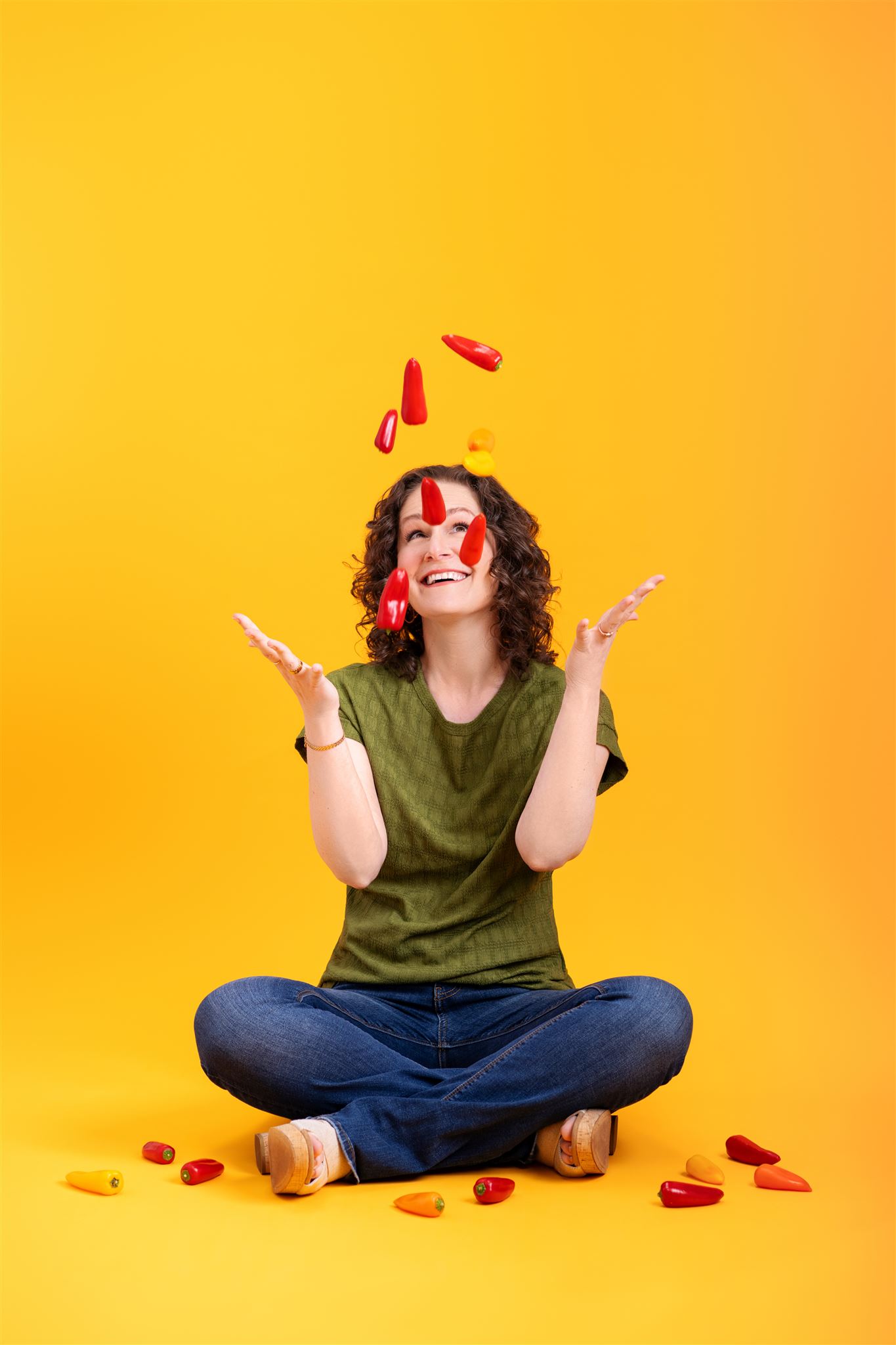 Smiling woman in front of a bright yellow background, tossing chile peppers into the air with several scattered on the ground—representing the playful spirit of Gringo Language Academy.