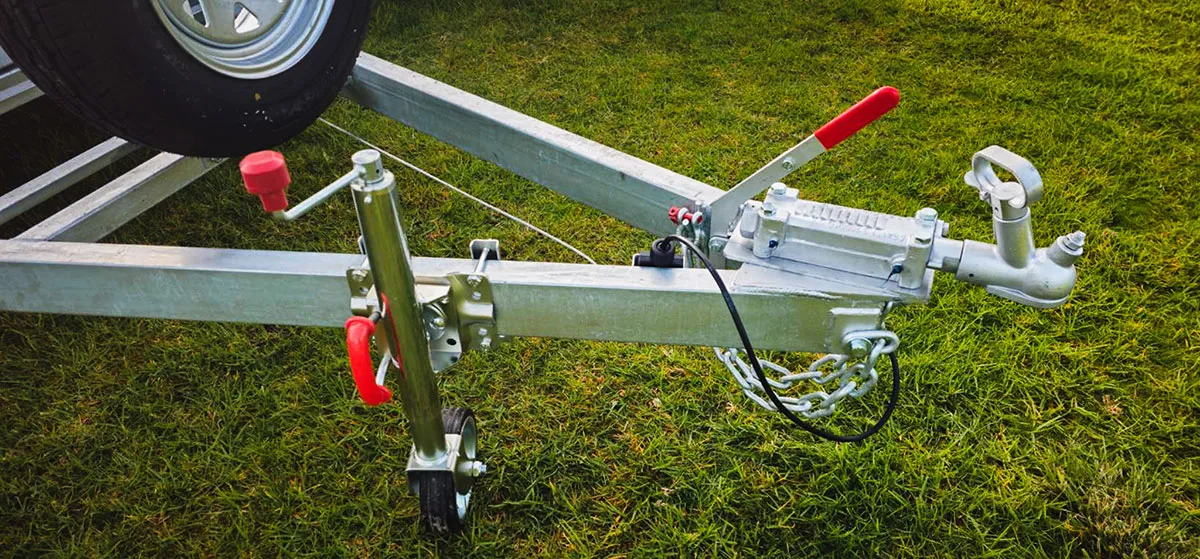 Close-up of a metal trailer hitch assembly with red handle, safety chains, and a small wheel on grass.