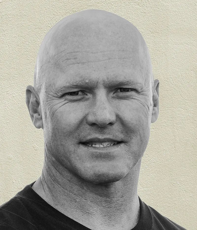 Close-up portrait of a man wearing a dark shirt against a textured light wall.