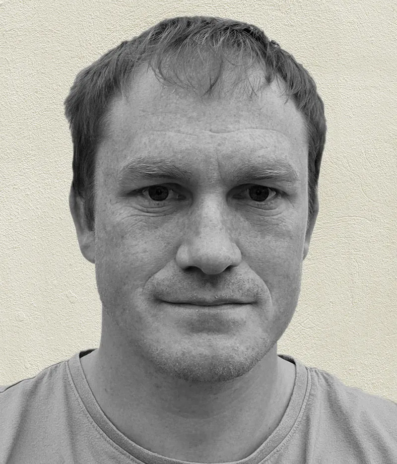 Close-up portrait of a man with short hair wearing a t-shirt against a textured light wall.