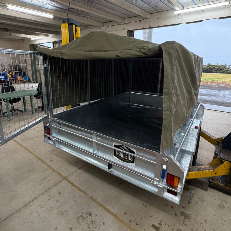 Metal trailer with mesh sides and green canvas cover inside a workshop with open side gate.