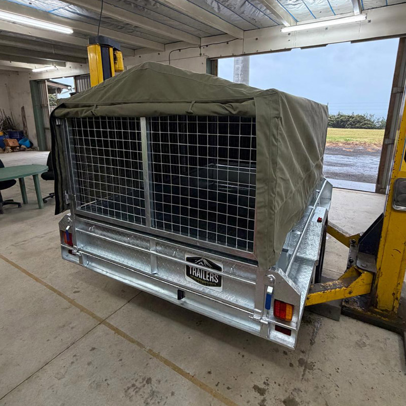 Metal trailer with a green canvas cover inside a workshop with yellow lifting equipment nearby.