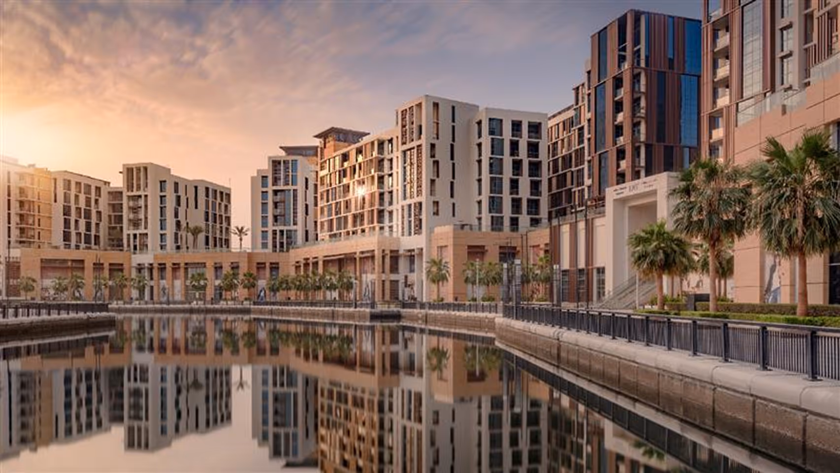 Modern residential buildings with palm trees lining a waterfront promenade reflected in calm water at sunset.