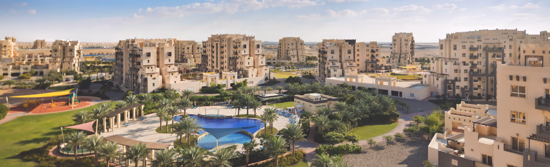 Residential complex with beige apartment buildings surrounding a central pool, playground, and landscaped garden area under a partly cloudy sky.