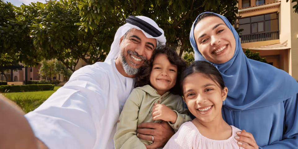 Smiling Middle Eastern family of four taking a selfie outdoors with greenery and buildings in the background.