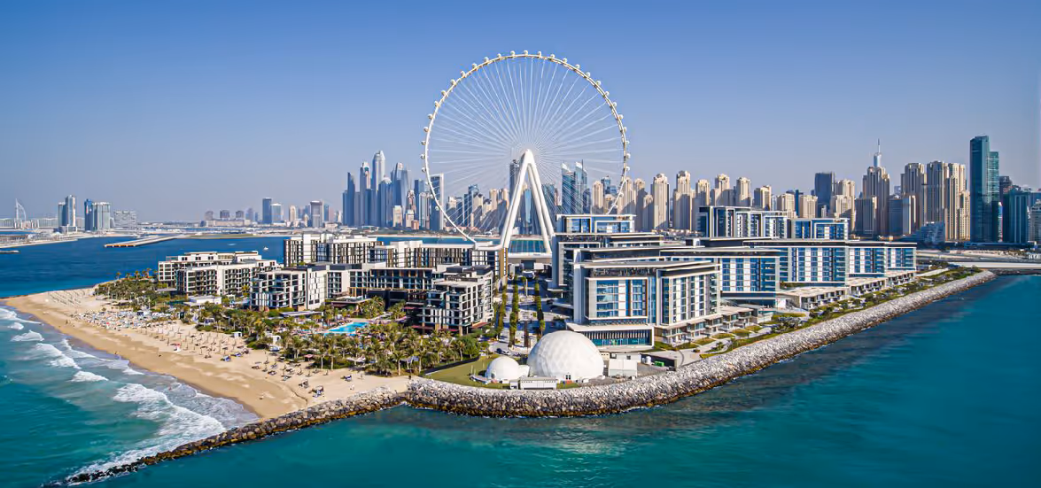 Aerial view of an island resort with modern buildings, a large Ferris wheel, and a city skyline in the background under clear blue sky.