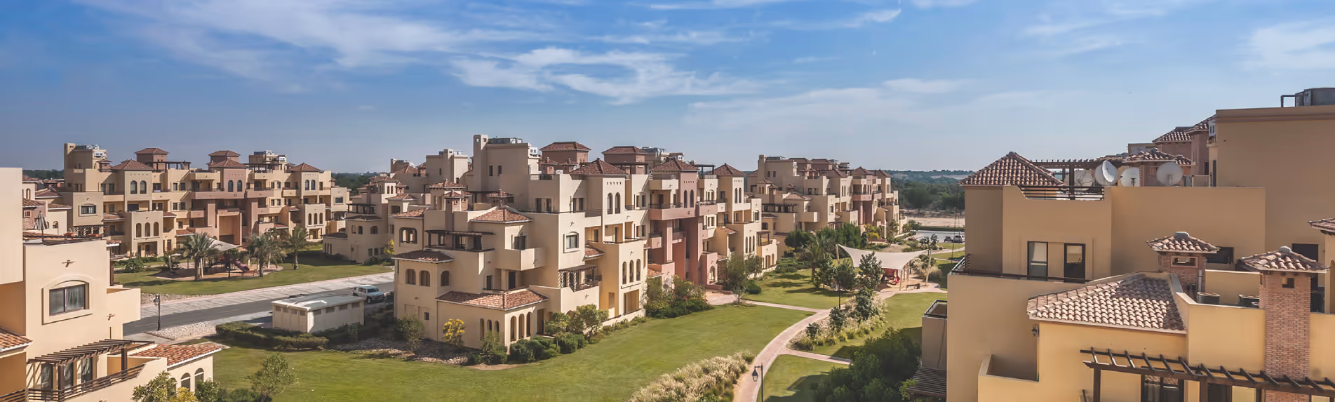 Residential community with Mediterranean-style buildings featuring terracotta roofs, surrounded by green lawns and pathways under a partly cloudy sky.