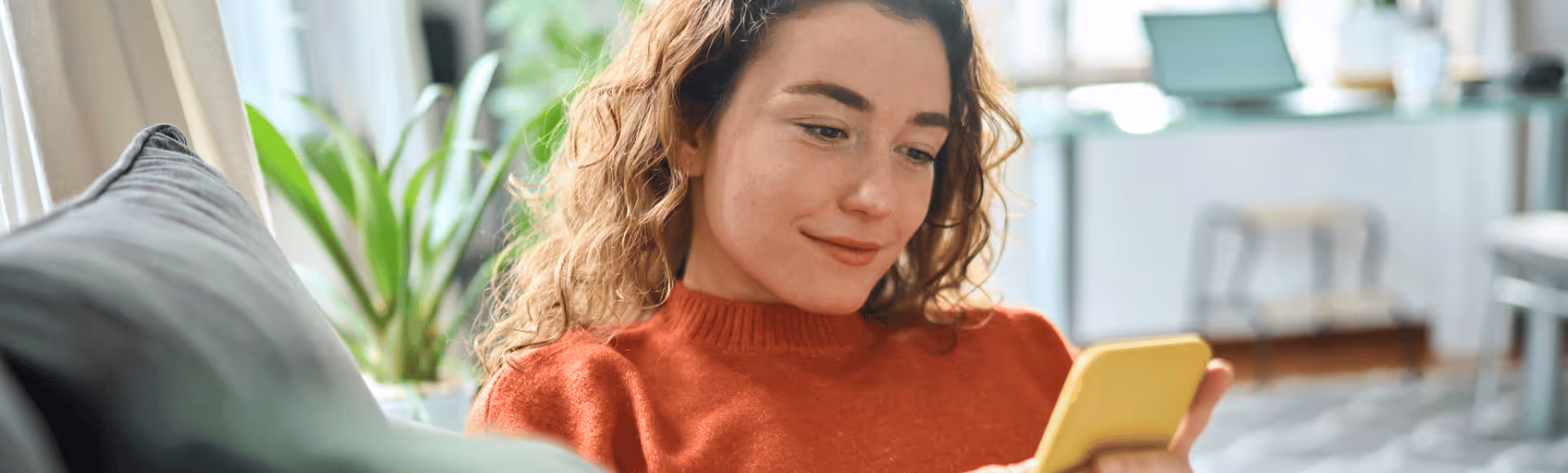 Young woman with curly hair wearing an orange sweater, smiling and looking at a yellow smartphone while sitting on a couch.