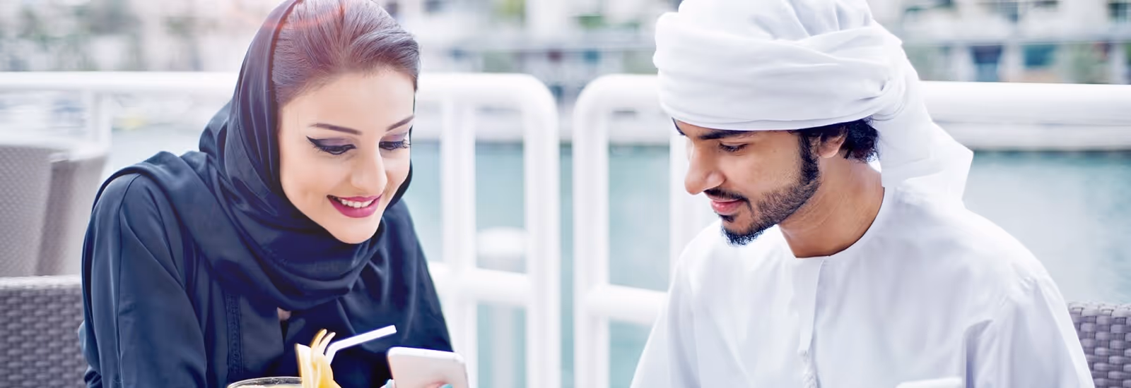 Smiling Middle Eastern woman in a black hijab and a man in white traditional dress looking at a smartphone together outdoors.