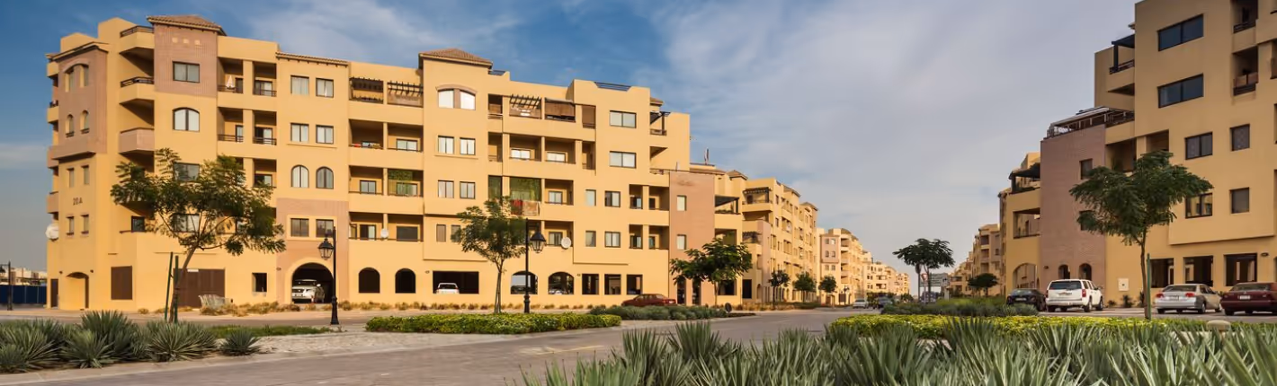 Wide street flanked by beige multi-story apartment buildings with trees and green plants under a partly cloudy sky.