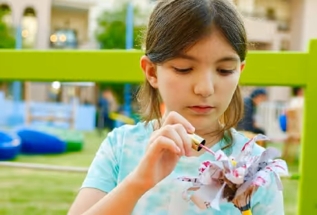 Young girl painting a flower craft outdoors on a green bench.