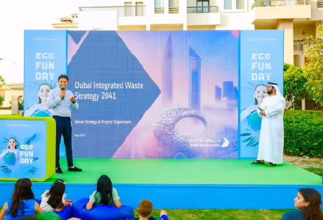 A man giving a presentation on Dubai Integrated Waste Strategy 2041 on an outdoor stage with audience seated on grass.