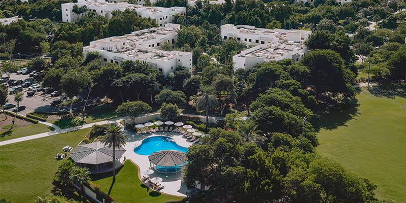 Aerial view of a residential area with white buildings, surrounded by dense green trees, a swimming pool, and a landscaped lawn.