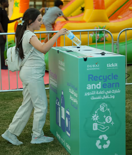 A young girl with a white flower in her hair recycling a plastic bottle in a green 'Recycle and earn' bin at an outdoor event with an inflatable slide in the background.