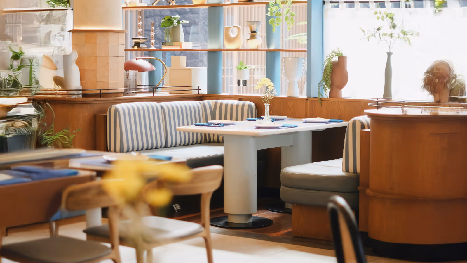 Cozy restaurant corner with striped cushioned booth seating, a white table set with plates and napkins, and decorative vases on shelves and window sills.