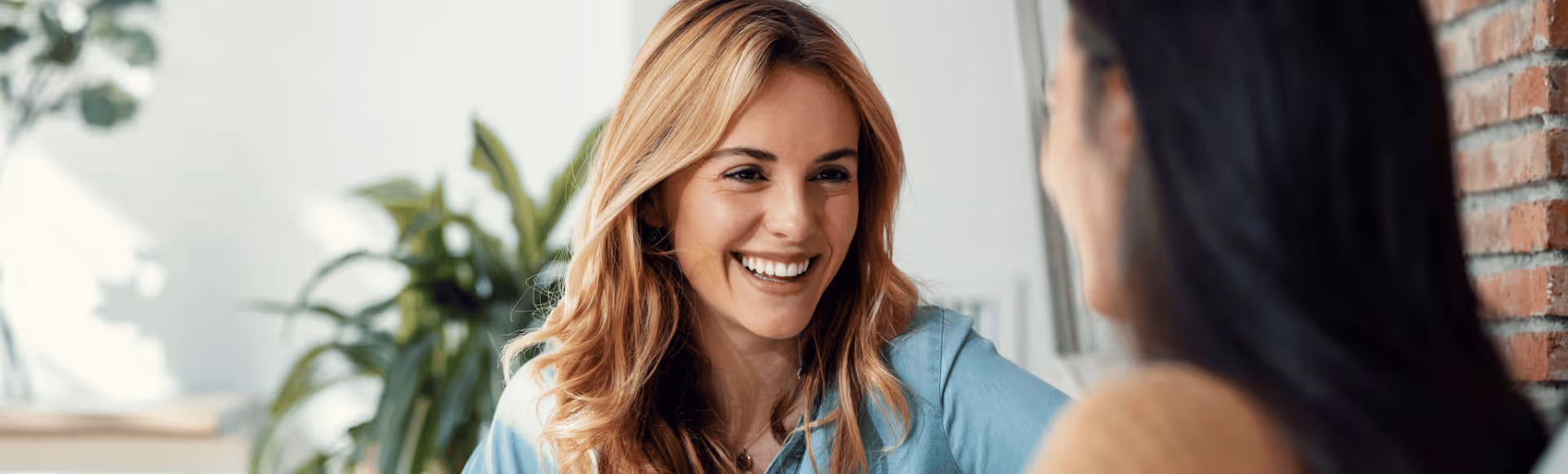 Smiling woman with blonde hair talking to another person in a bright indoor setting with plants and a brick wall.