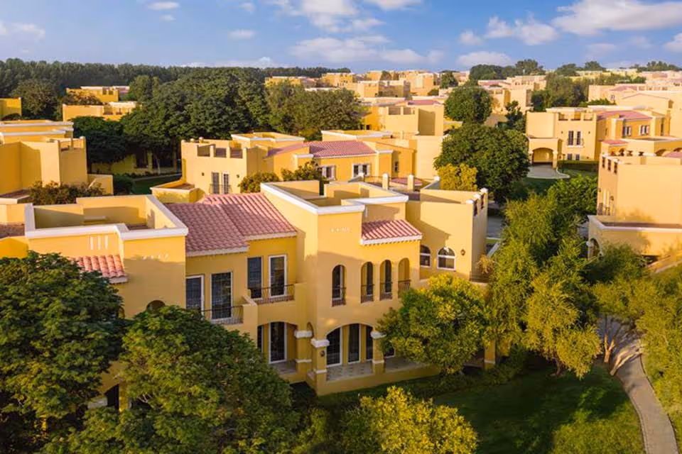 Aerial view of a residential neighborhood with yellow Mediterranean-style houses and green trees under a blue sky with scattered clouds.