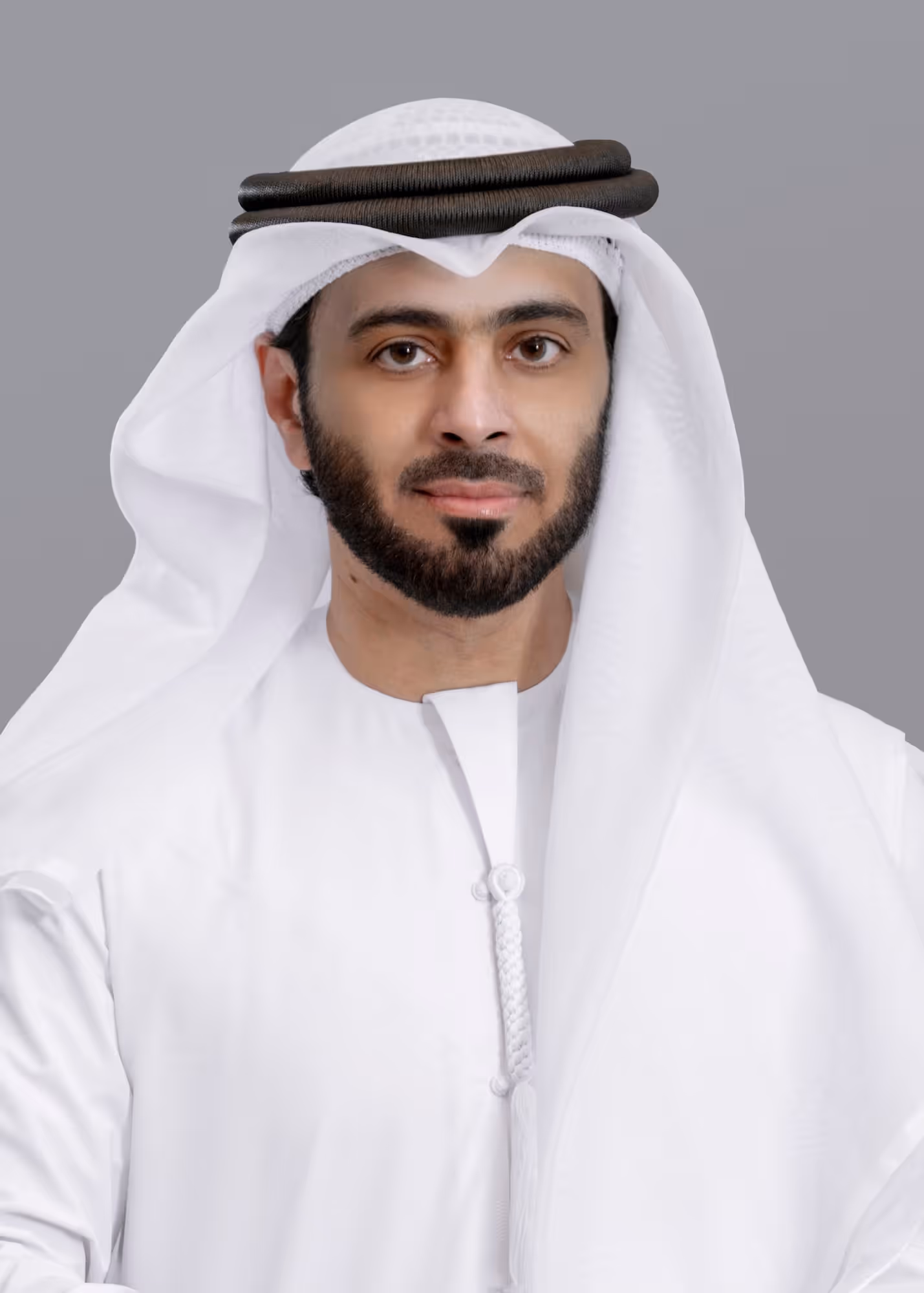 Portrait of a man wearing a white kandura and traditional Emirati headgear with a black agal on a gray background.