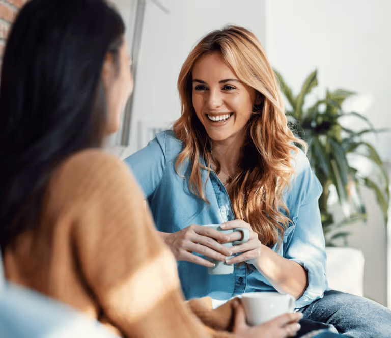 Two women sitting and smiling while holding coffee mugs during a conversation indoors.