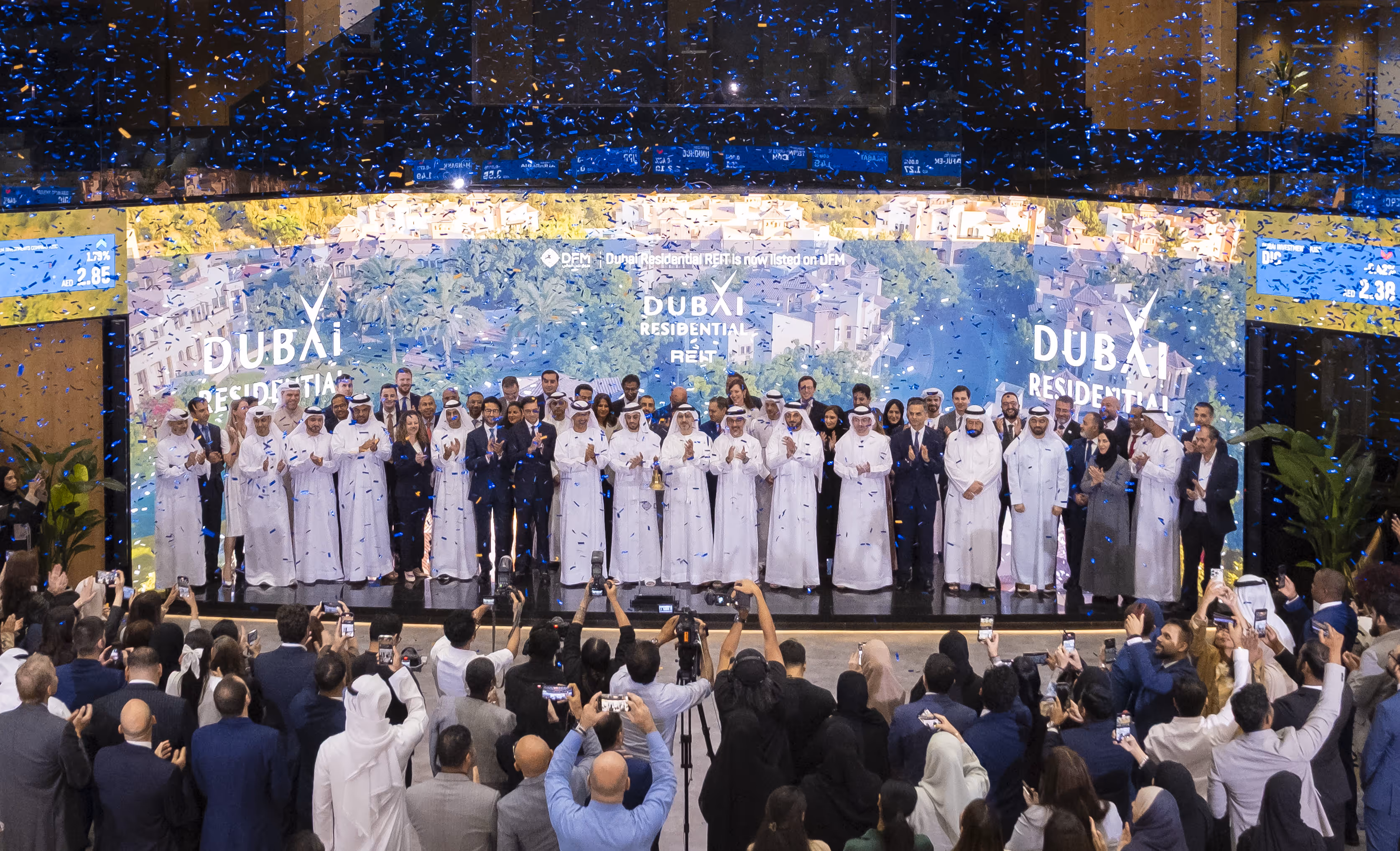 Group of people dressed in traditional Emirati attire and formal suits celebrating on stage with blue confetti falling, Dubai Residential REIT branding in background.
