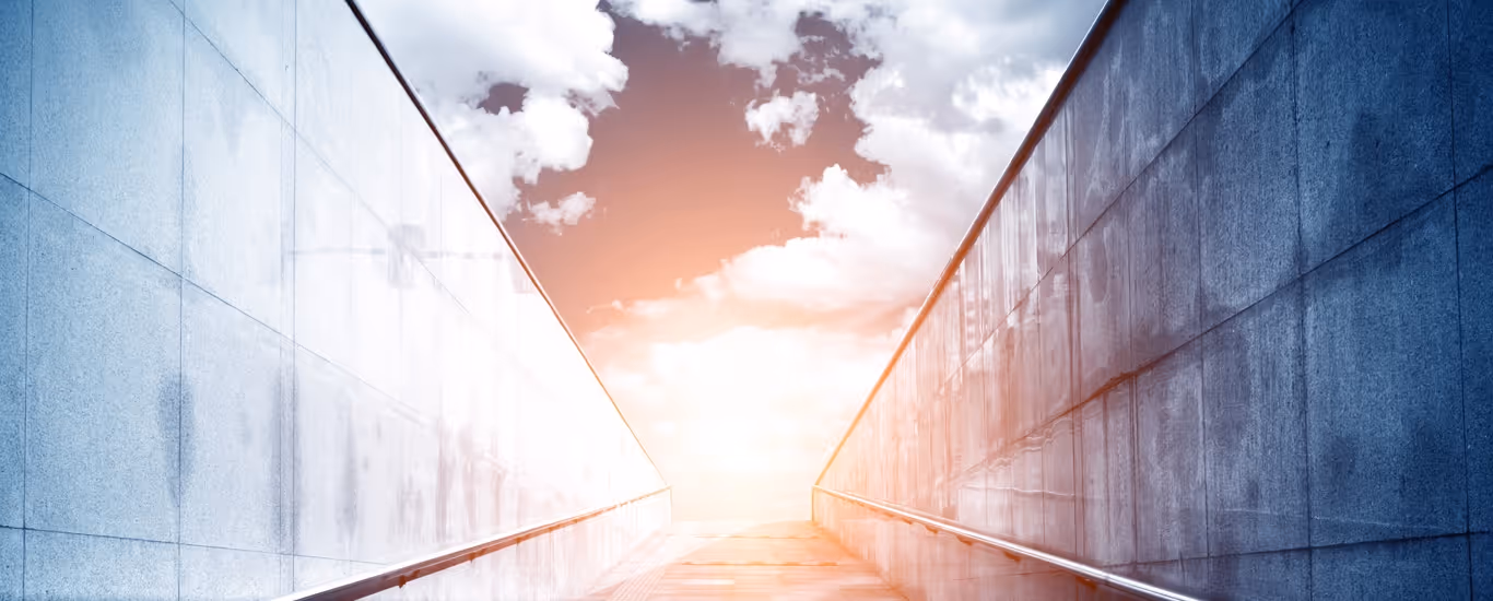 Stairway between two reflective stone walls leading up to a bright sky with scattered clouds and sunlight.