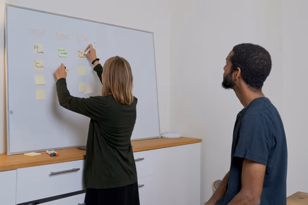 two people sit at a whiteboard looking at a task list