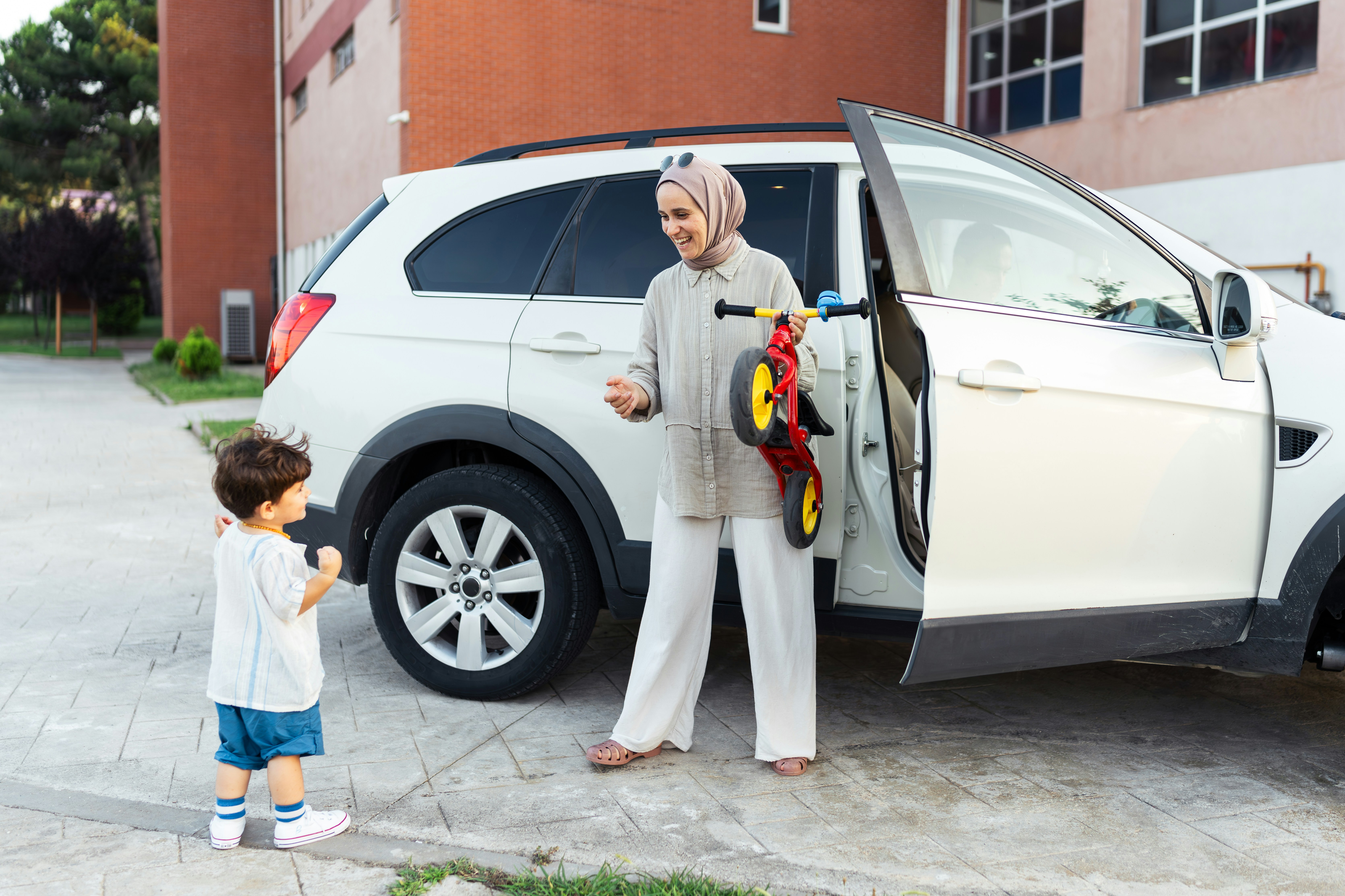 Woman in hijab holding a colourful tricycle next to a car with a toddler in front of her.