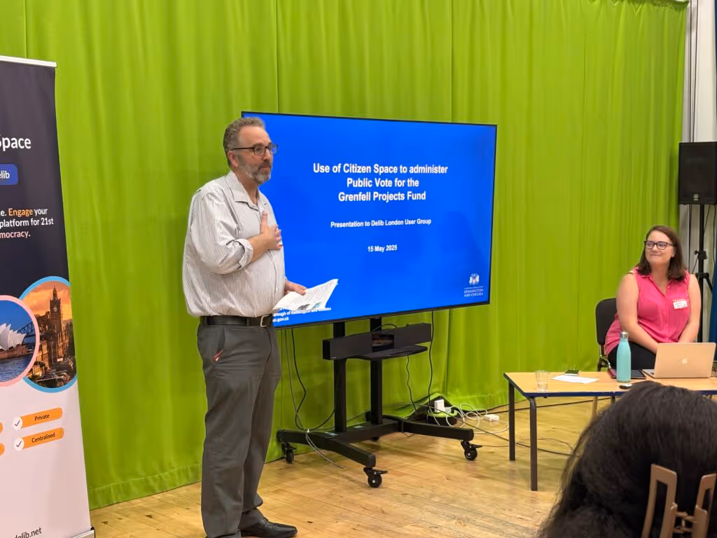 Gary Wilson, a man with salt-and-pepper coloured hair and beard wearing grey trousers and a pale shirt stands in front of a large screen to his left. The screen displays a slide from his presentation about Kensington and Chelsea Council's use of Citizen Space to administer the public vote fro the Grenfell Projects Fund.