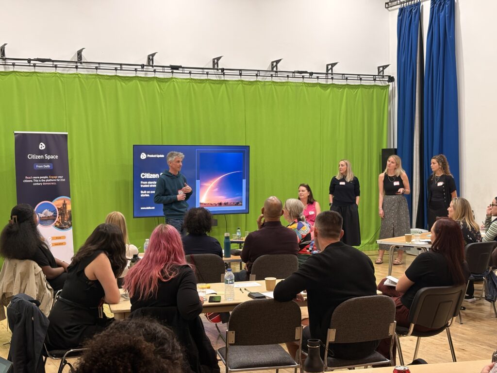 Andy Parkhouse, with grey heair and wearing a green hooded sweatshirt, stands in front of a large screen and addresses a seated audience. Looking on from Andy's left are Jenni Weech, wearing a pink top, Louise Cato, wearing all black, Jessie Ashmore, wearing a black top and grey patterned skirt, and Eli Watson, also dressed all in black.