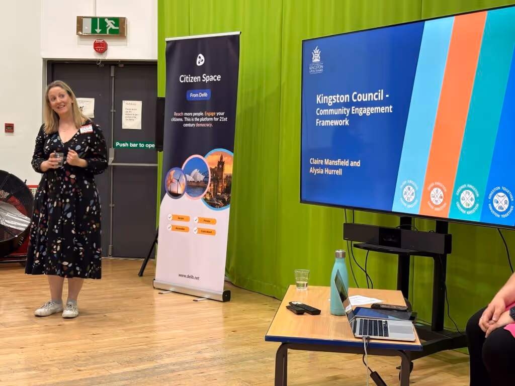 Claire Mansfield, a woman with long blonde hair wearing a dark patterned dress, stands looking at a large screen to her left. The screen displays a slide from her presentation about Kingston Council's Community Engagement Framework.